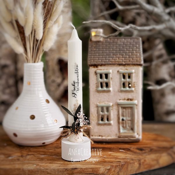 Weiße Weihnachtskerze mit dem Schriftzug Frohe Weihnachten, kleinem Weihnachtsbaum aus Holz, grünen Blättern und zarten Blüten.