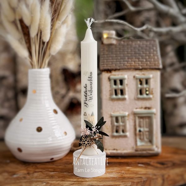 Weiße Weihnachtskerze mit dem Schriftzug Frohe Weihnachten, winterlichen Häusern, Holzschneeflocke und grünen Blättern.